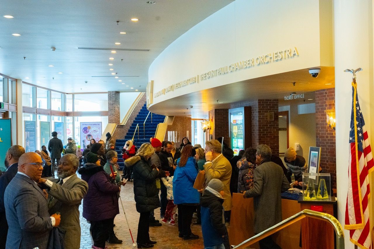 Crowd of diverse people wearing winter clothing standing in line in a theater lobby with ticket counter, blue carpeted stairs, and an American flag on the right.